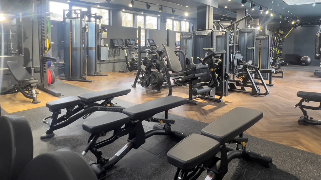 Big open gym with wooden floors and windows; bench press equipment in foreground