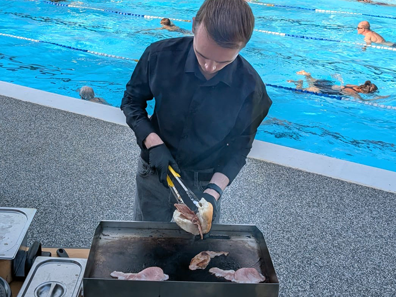 young man using tongs to put bacon in a roll above barbecue, swimming pool in background with swimmers