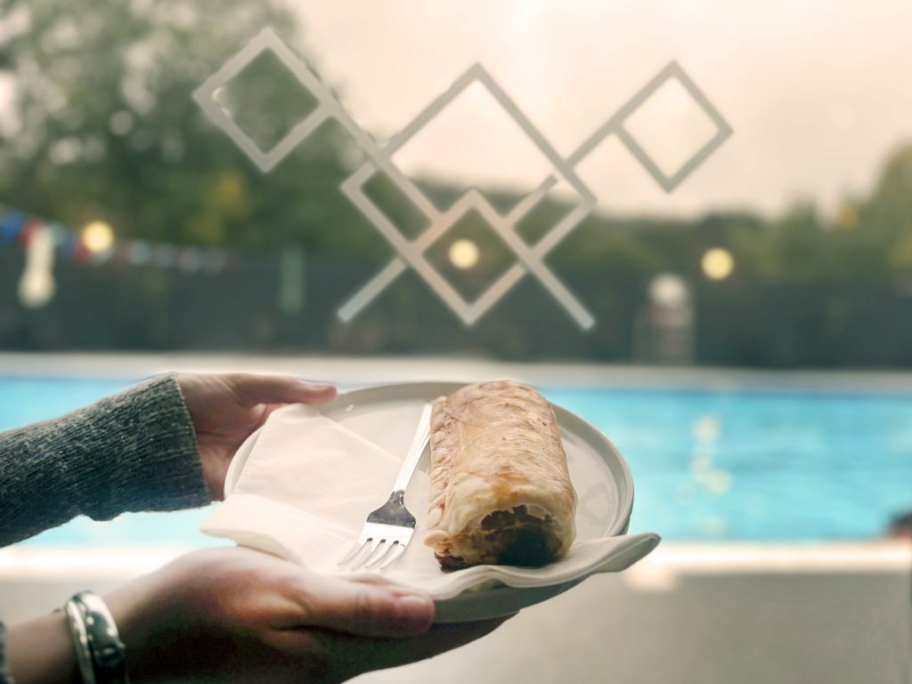 hands holding a plate with a suasge roll with swimming pool in background